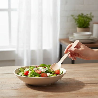 Hand holding a sustainable Food Packaging Direct wooden spork over a fresh salad bowl in a bright kitchen setting.